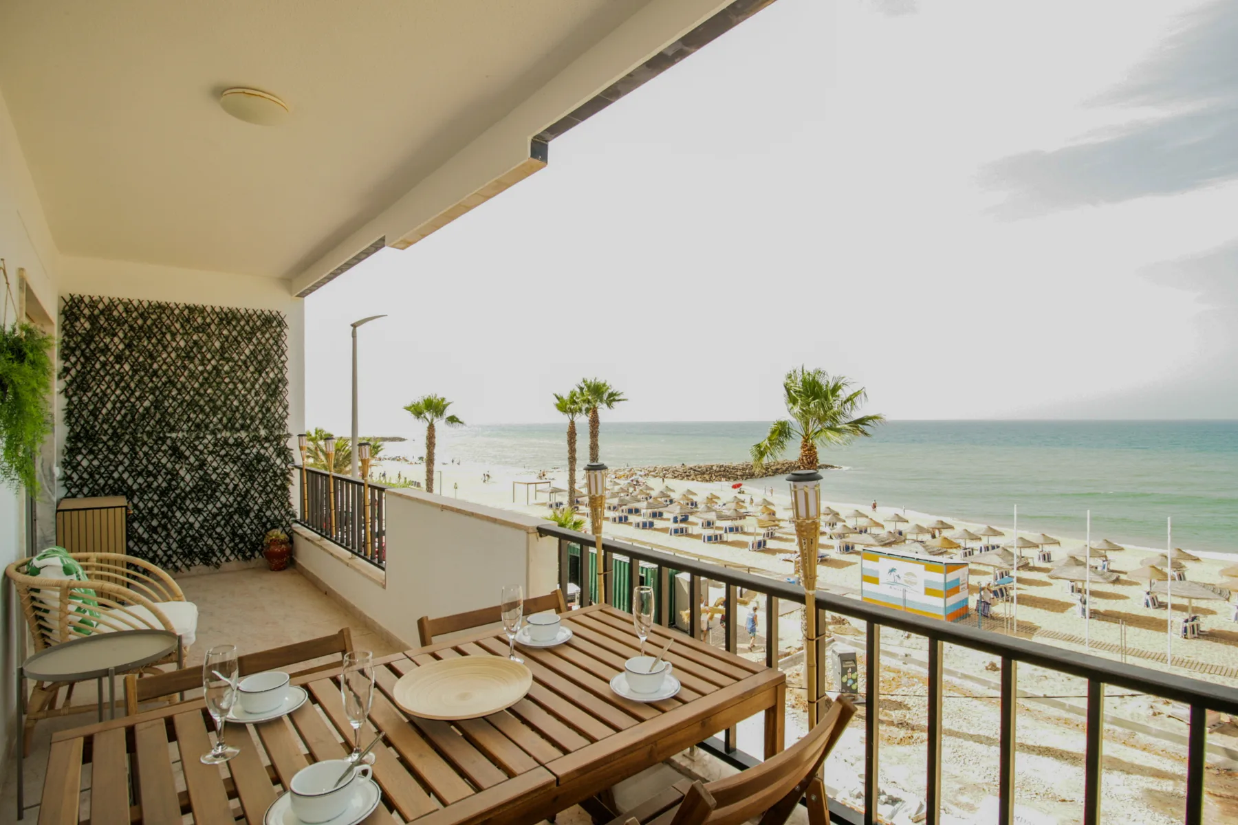 A balcony dining area overlooks a beach with rows of umbrellas and the sea.