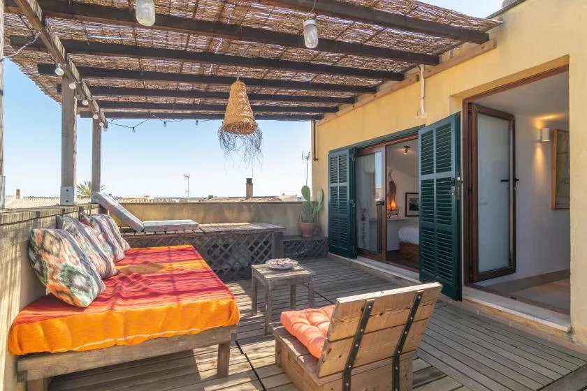 Una terraza soleada con diván exterior y asientos de madera bajo una pérgola sombreada.