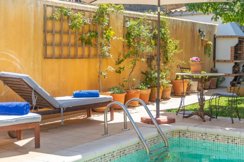 Un patio soleado con asientos junto a la piscina, plantas en macetas y una pequeña mesa al aire libre.