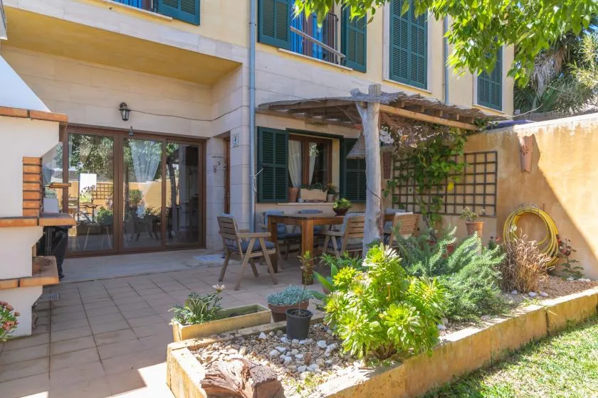 Un patio soleado junto a una casa, con asientos al aire libre y un jardín plantado.