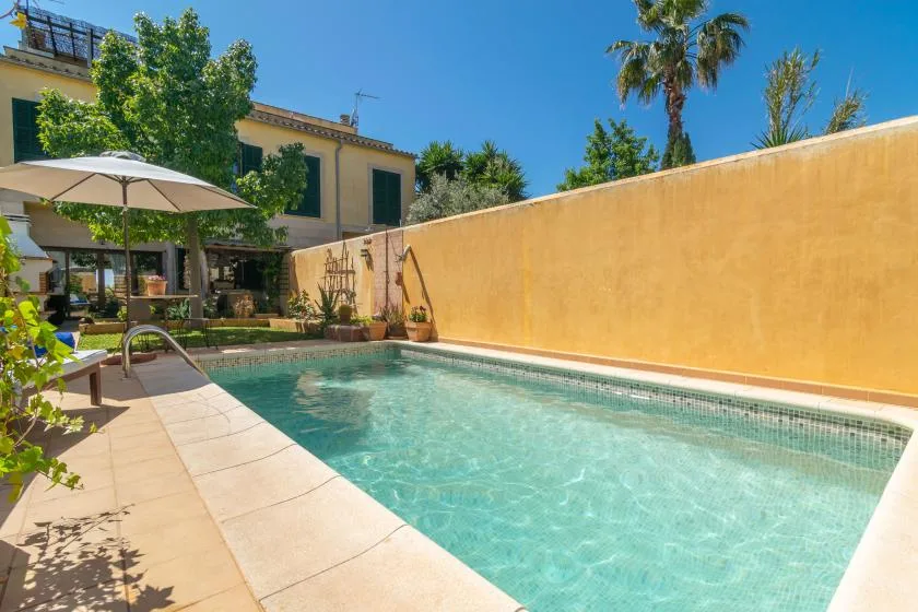 Una piscina junto a un patio soleado y un muro alto.