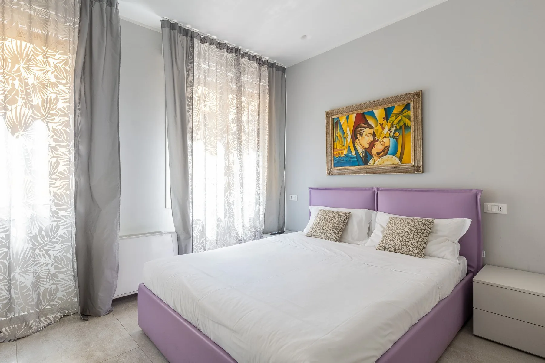 Un dormitorio luminoso con una cama morada, ropa de cama blanca y ventanas altas con cortinas.