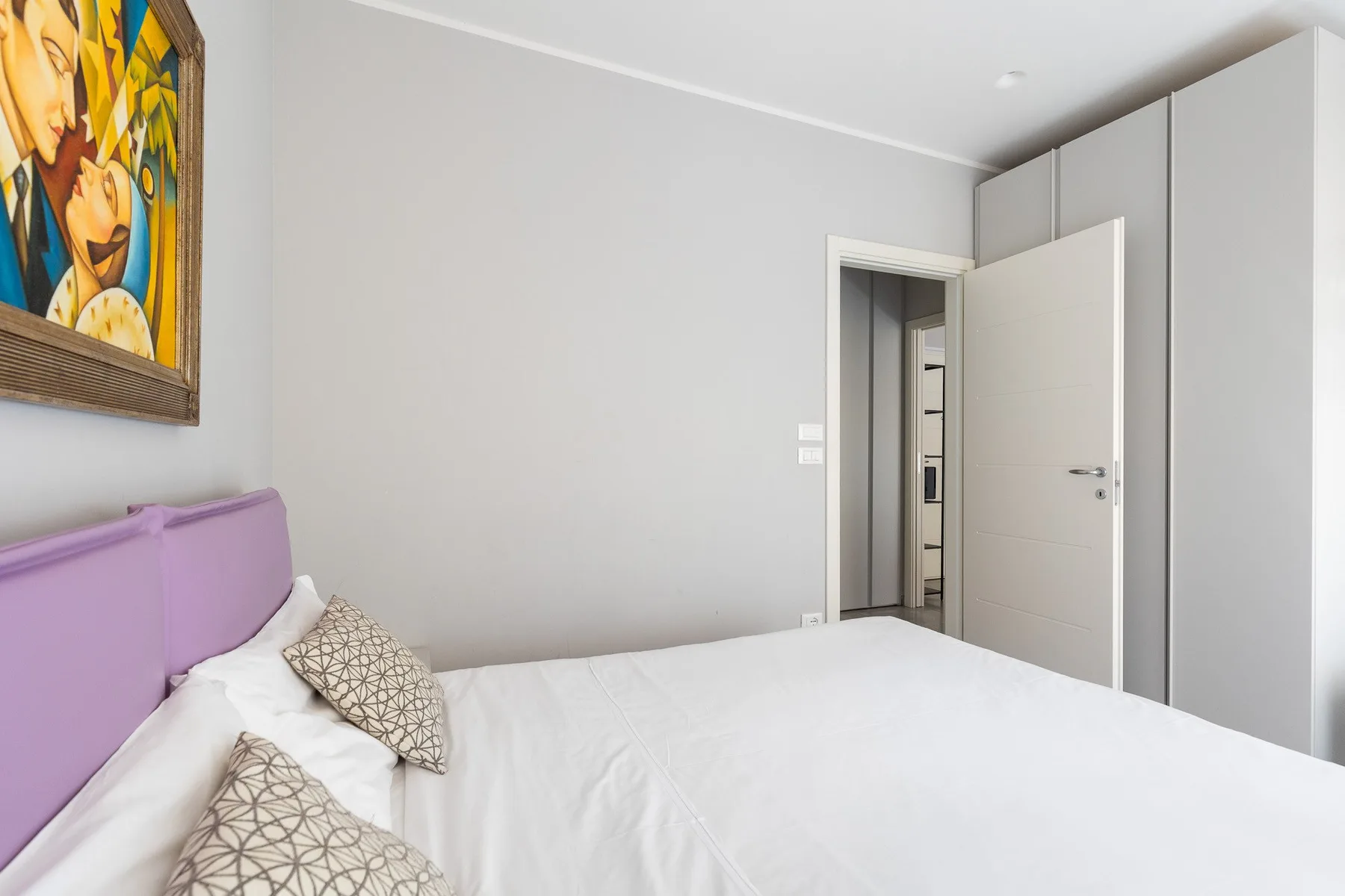 Un dormitorio con una cama blanca, un cabecero morado y una puerta abierta.