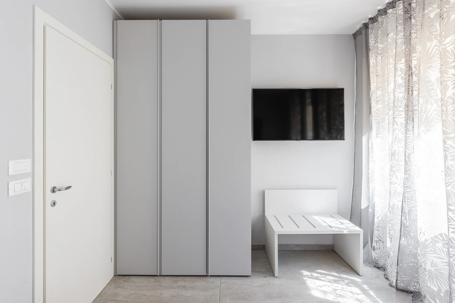 Una habitación luminosa con un armario alto, un televisor en la pared y una cama baja.