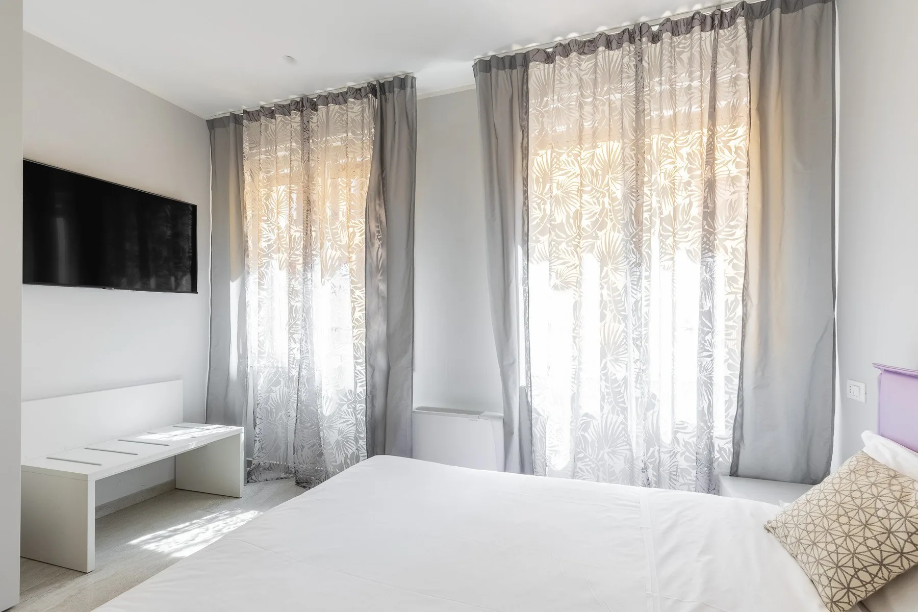 Un dormitorio luminoso con cama, televisor en la pared y ventanas altas con cortinas.