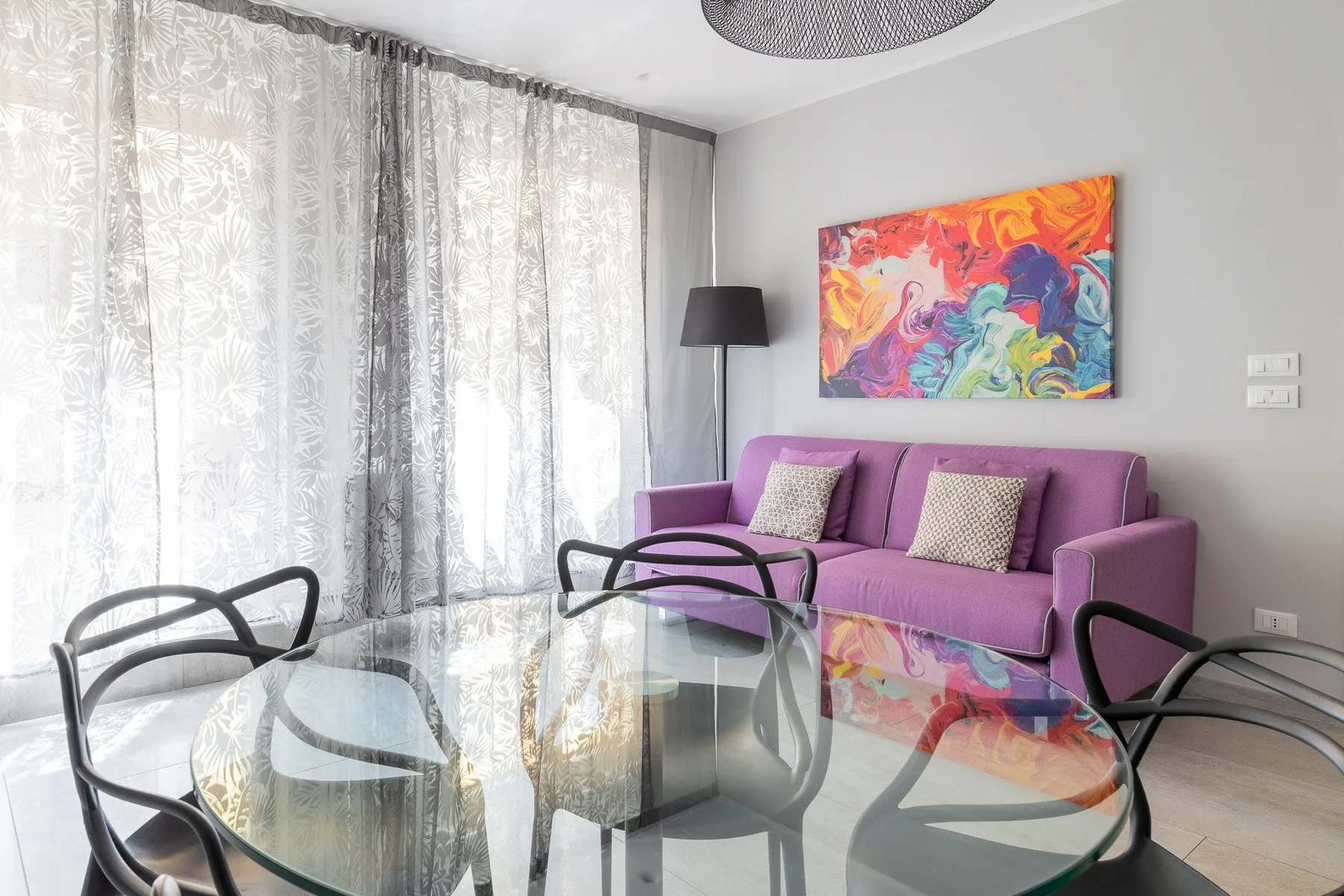 Una sala de estar con asientos morados, muebles de vidrio y arte colorido en la pared.