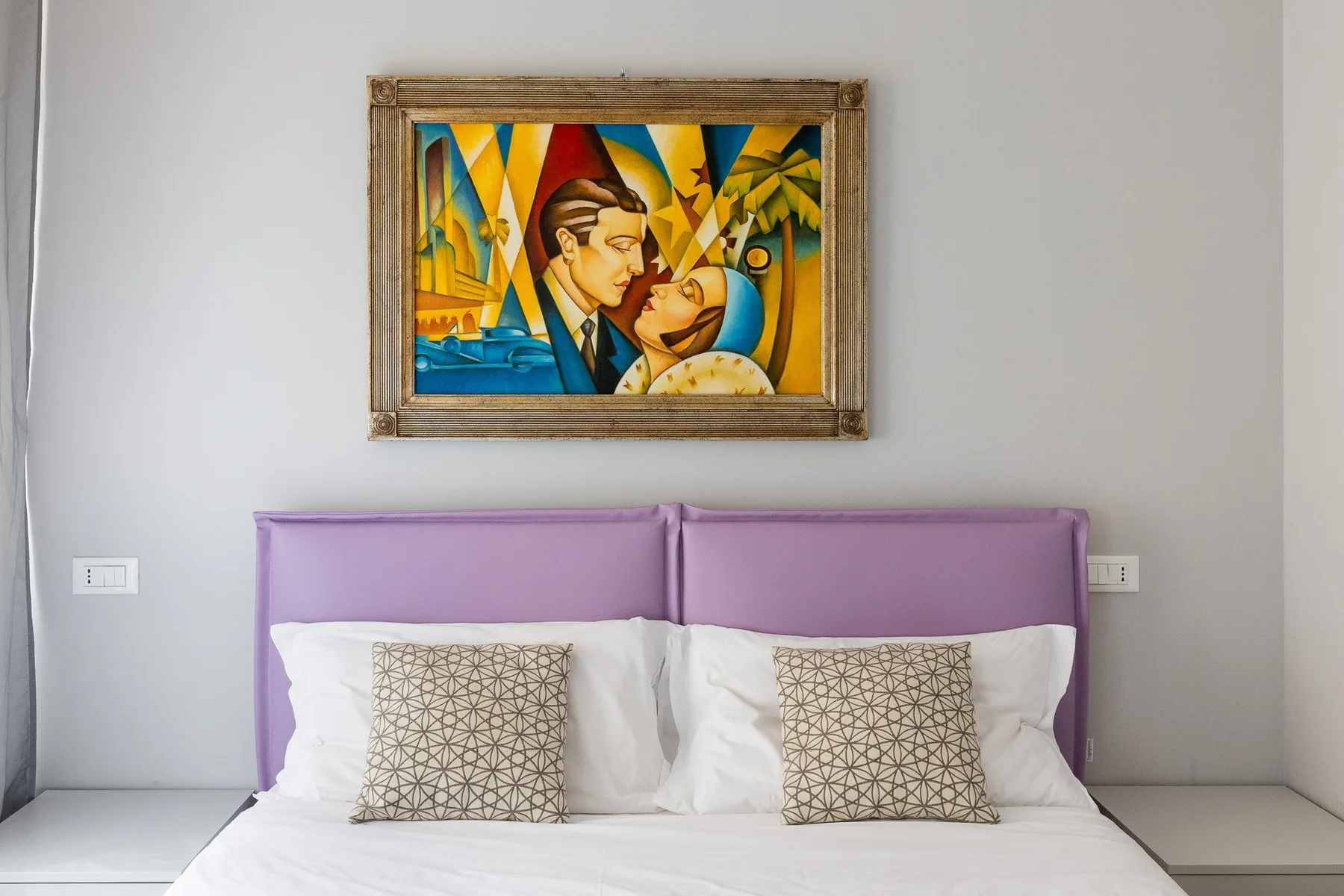 Un dormitorio con una cama, un cabecero tapizado morado y una colorida obra enmarcada.