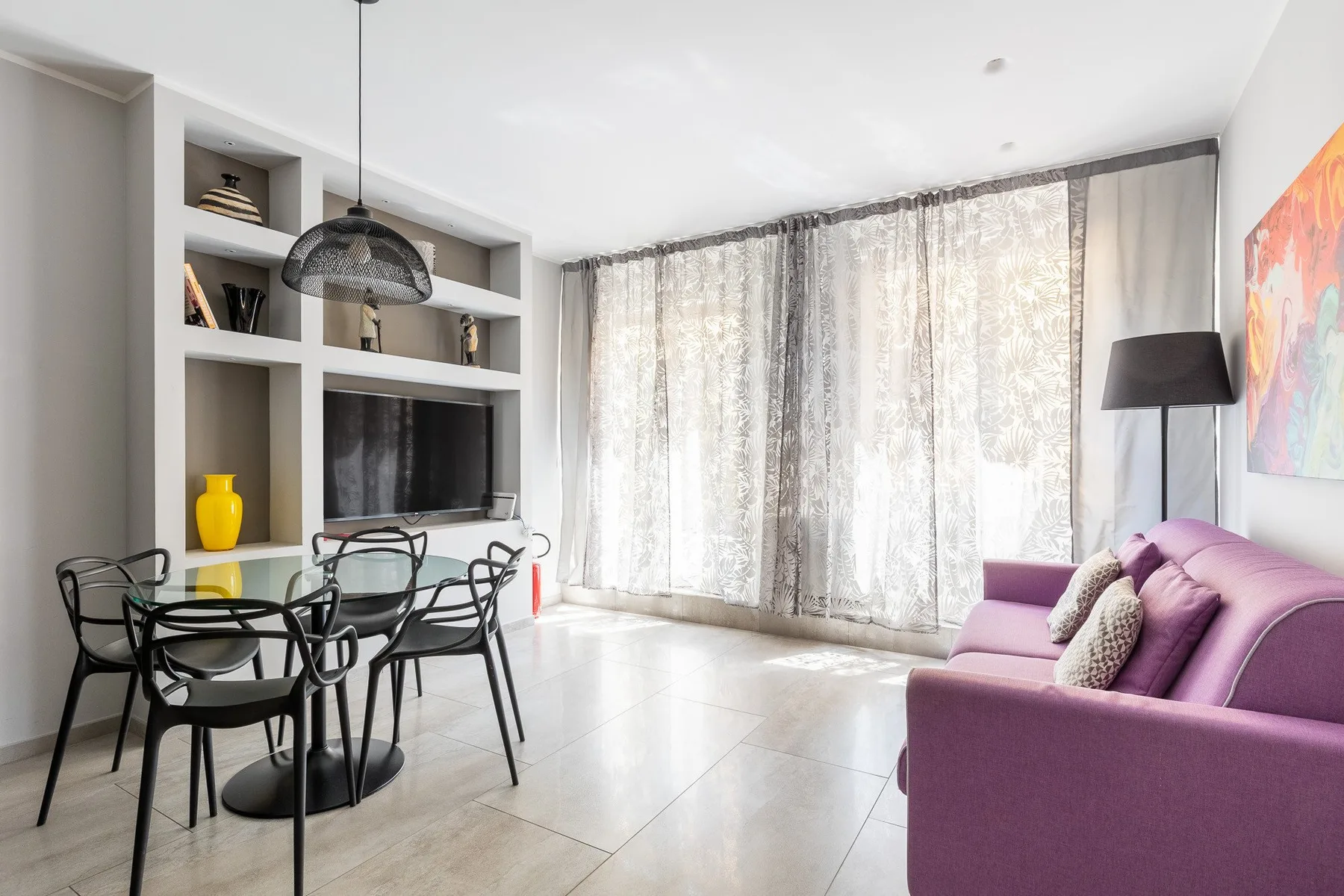 Una luminosa sala de estar y comedor con un sofá morado y una mesa junto a grandes ventanales.