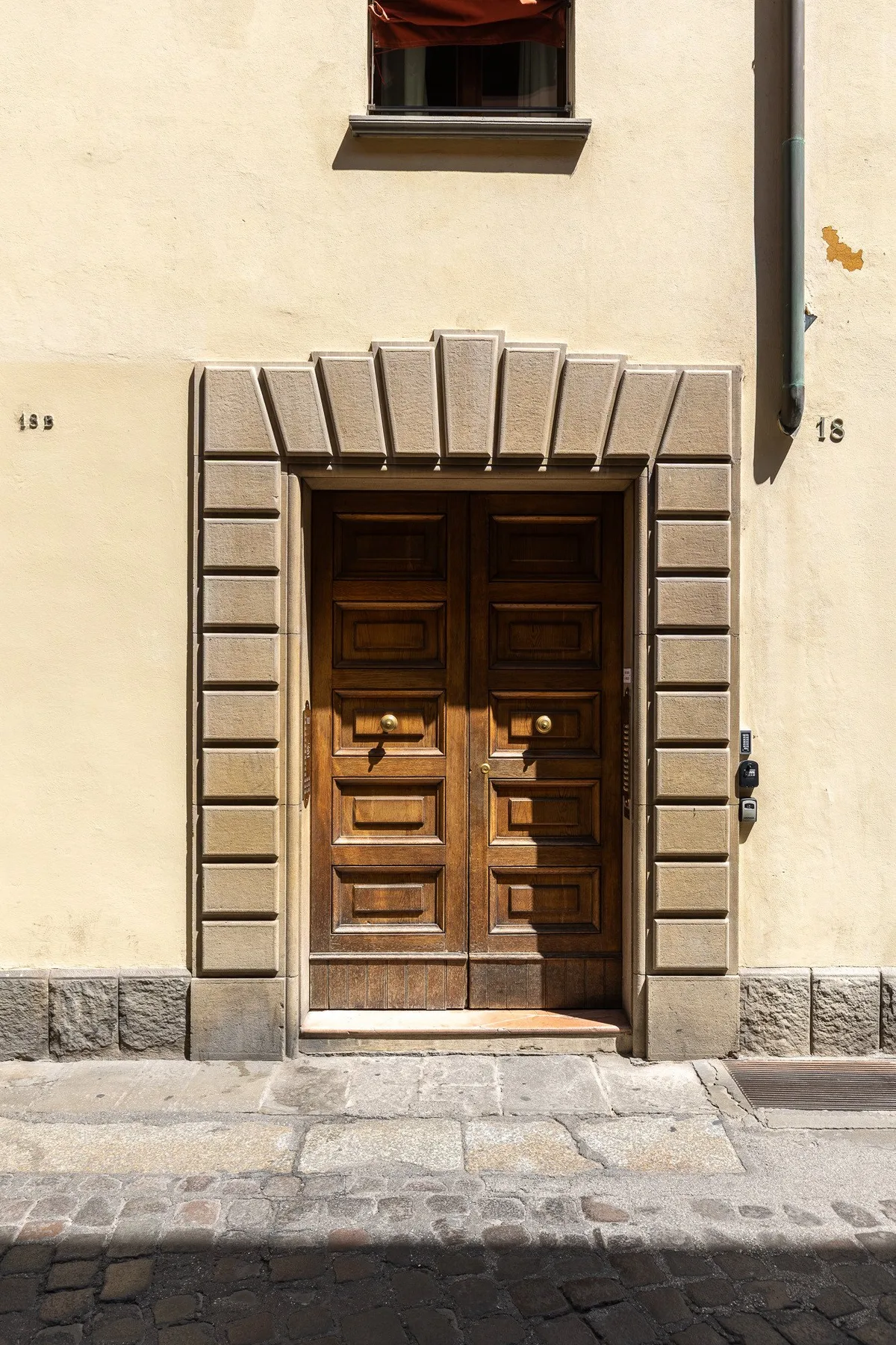 Una puerta doble de madera oscura está en una fachada clara, sobre una acera de piedra.