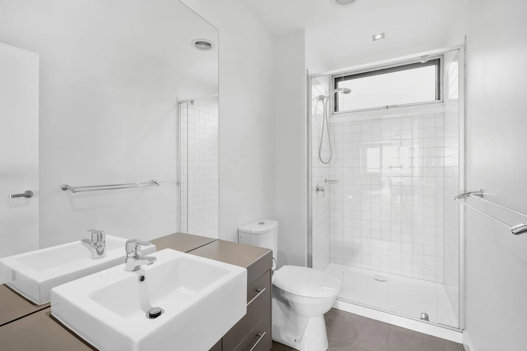 A bright bathroom with double sinks, a toilet, and a glass shower.