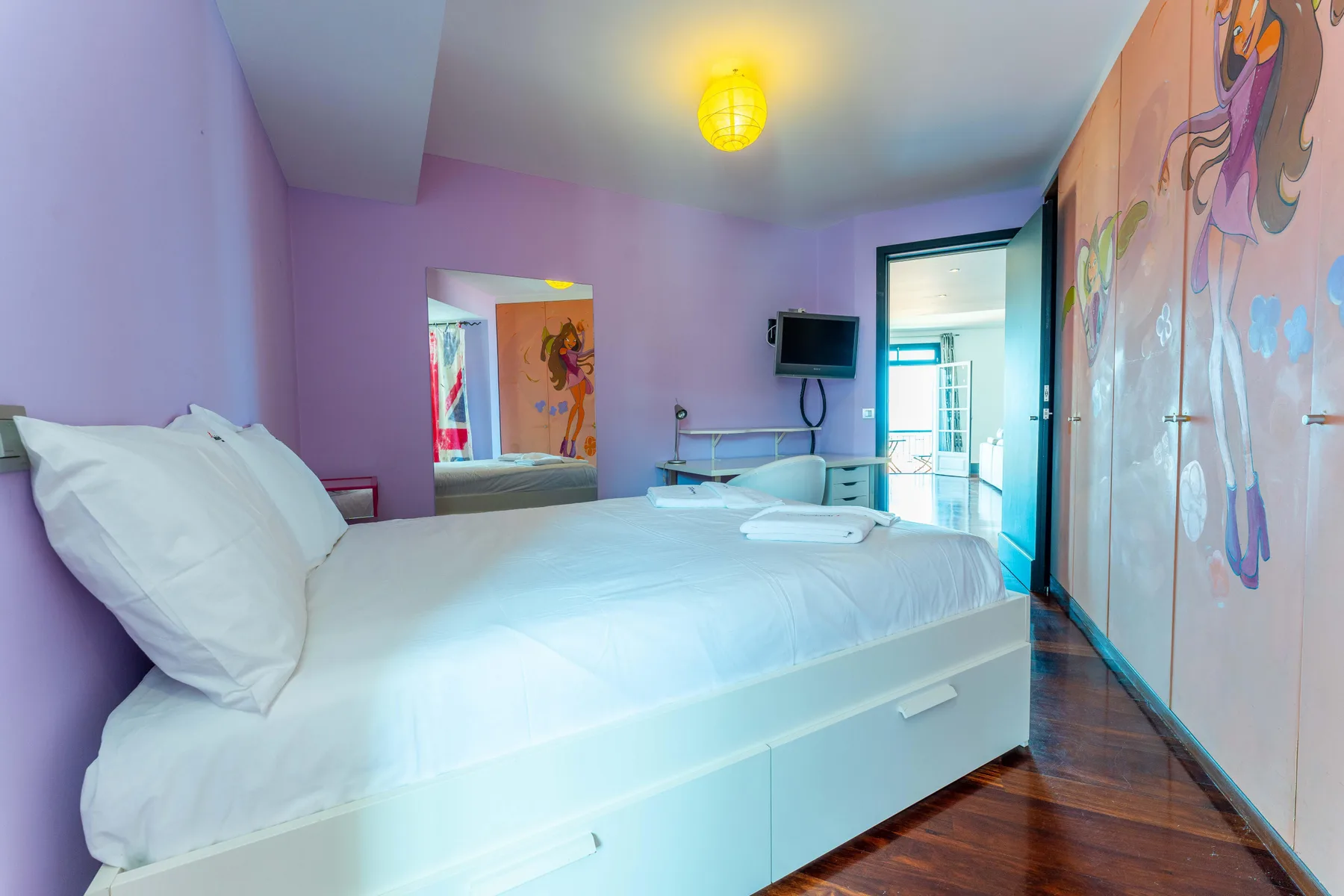 A bedroom with a large bed, colorful wall art, and a doorway opening onto a bright area.