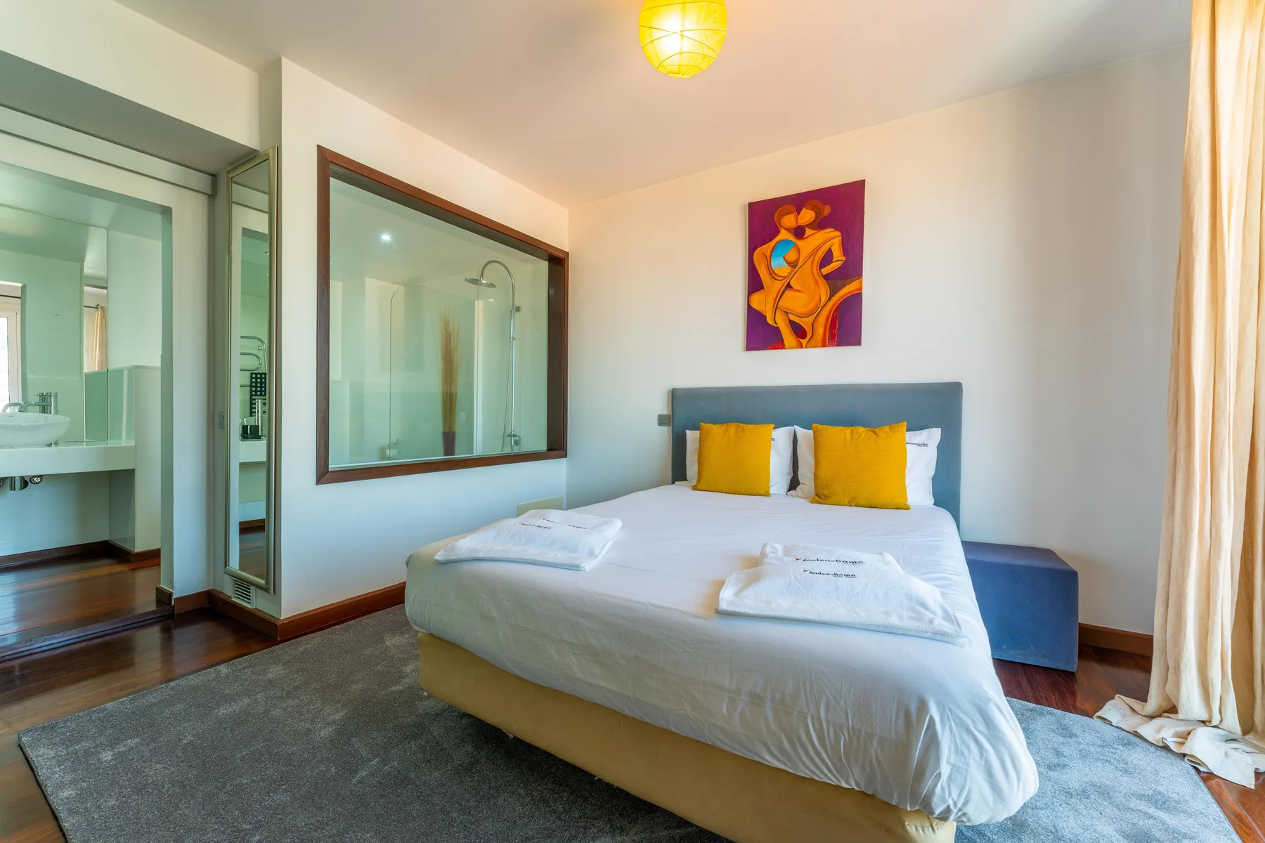 A bedroom with a neatly made bed, yellow pillows, and colorful artwork above the headboard.