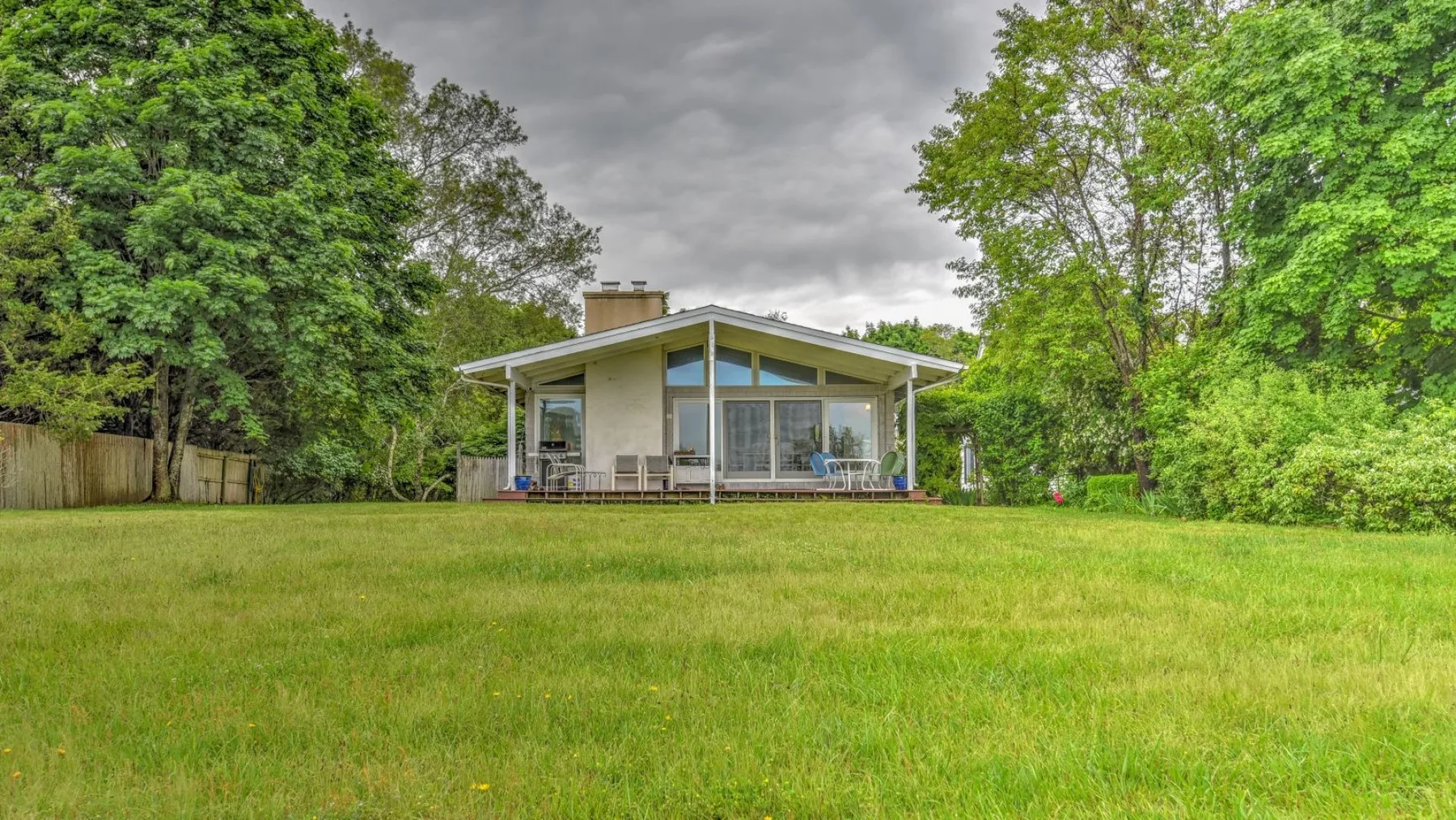 A modern house stands beyond a broad green lawn, surrounded by trees.