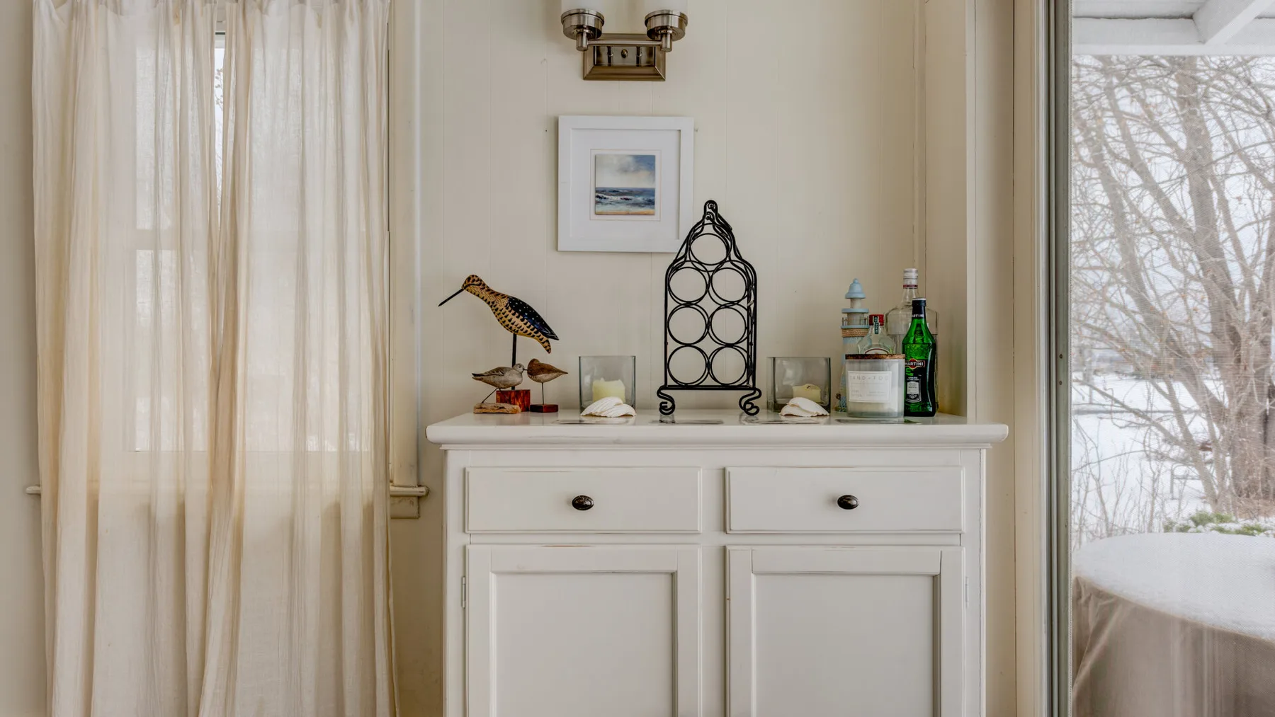 A white cabinet with decorative objects stands between curtained windows.