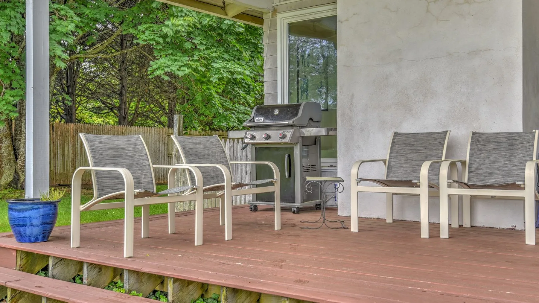 A covered outdoor deck has seating, a grill, and leafy trees beyond the railing.