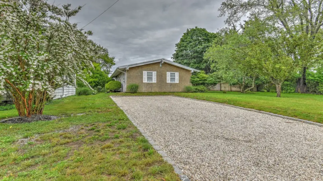 A single-story house stands at the end of a broad gravel driveway.