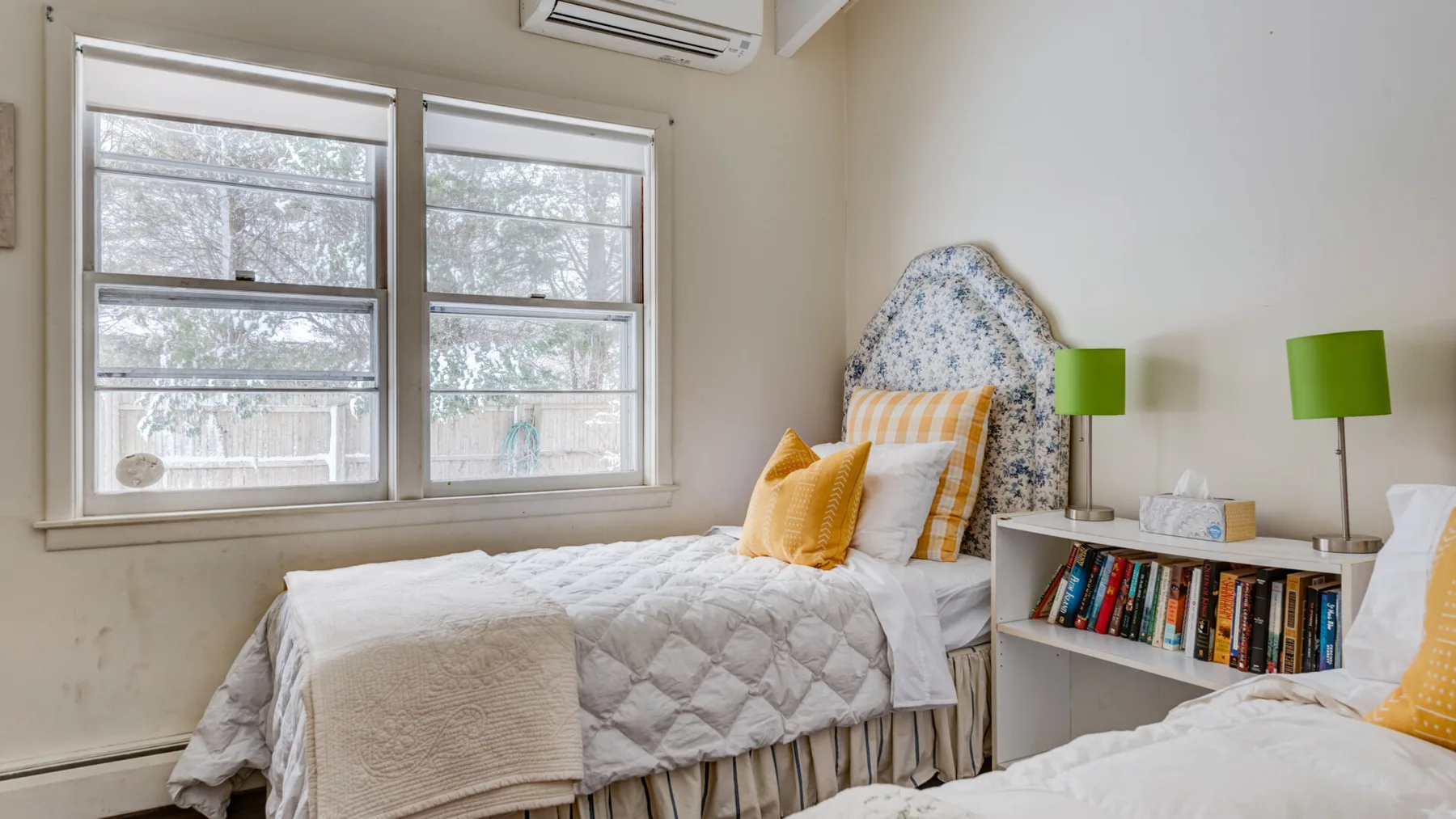 A bedroom with two beds, a bookshelf, and large windows.