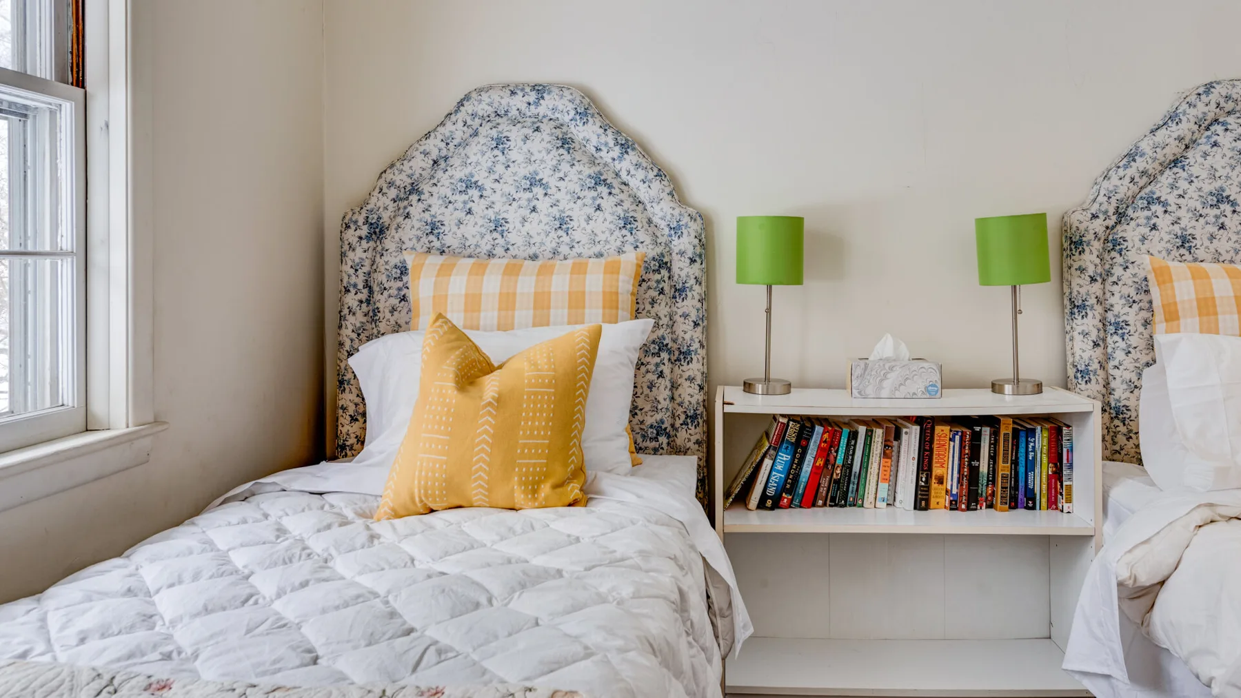 A bedroom with two single beds, colorful pillows, and a low bookshelf between them.