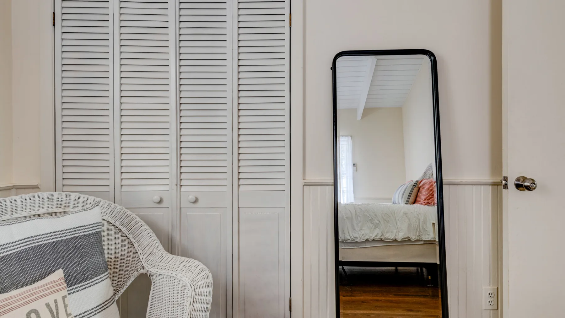 A bedroom with a bed reflected in a tall mirror beside louvered closet doors.
