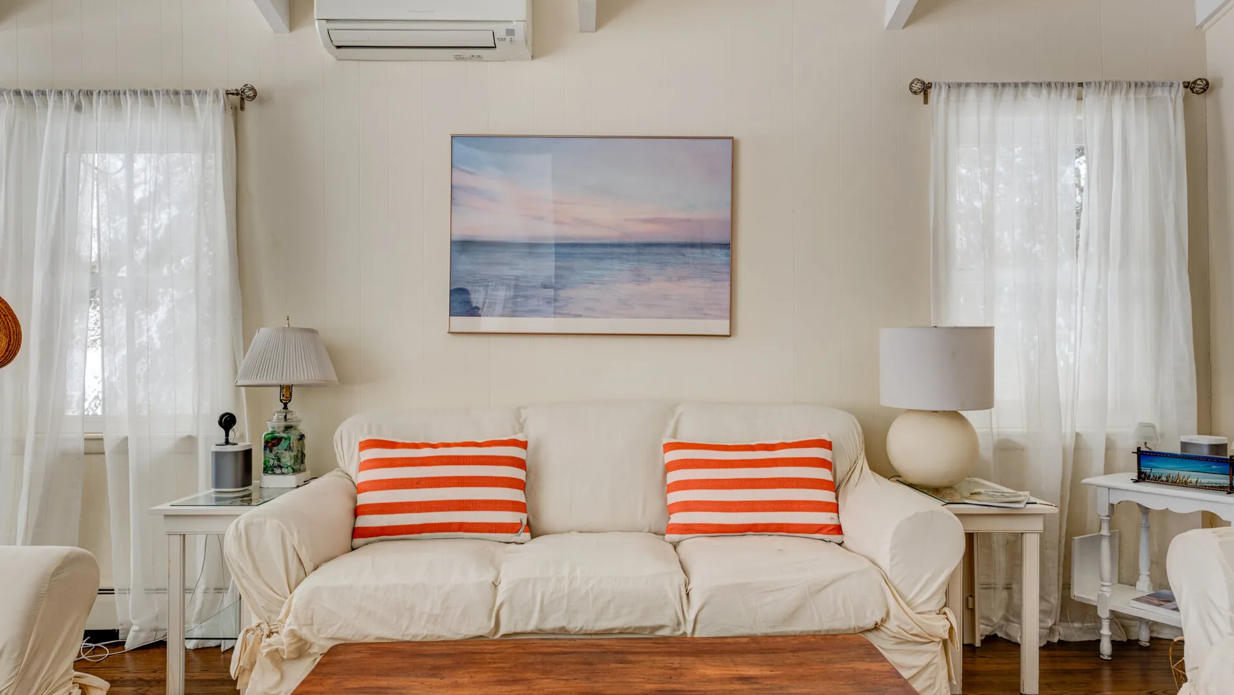 A bright living room with cream seating, striped cushions, and a framed seascape.