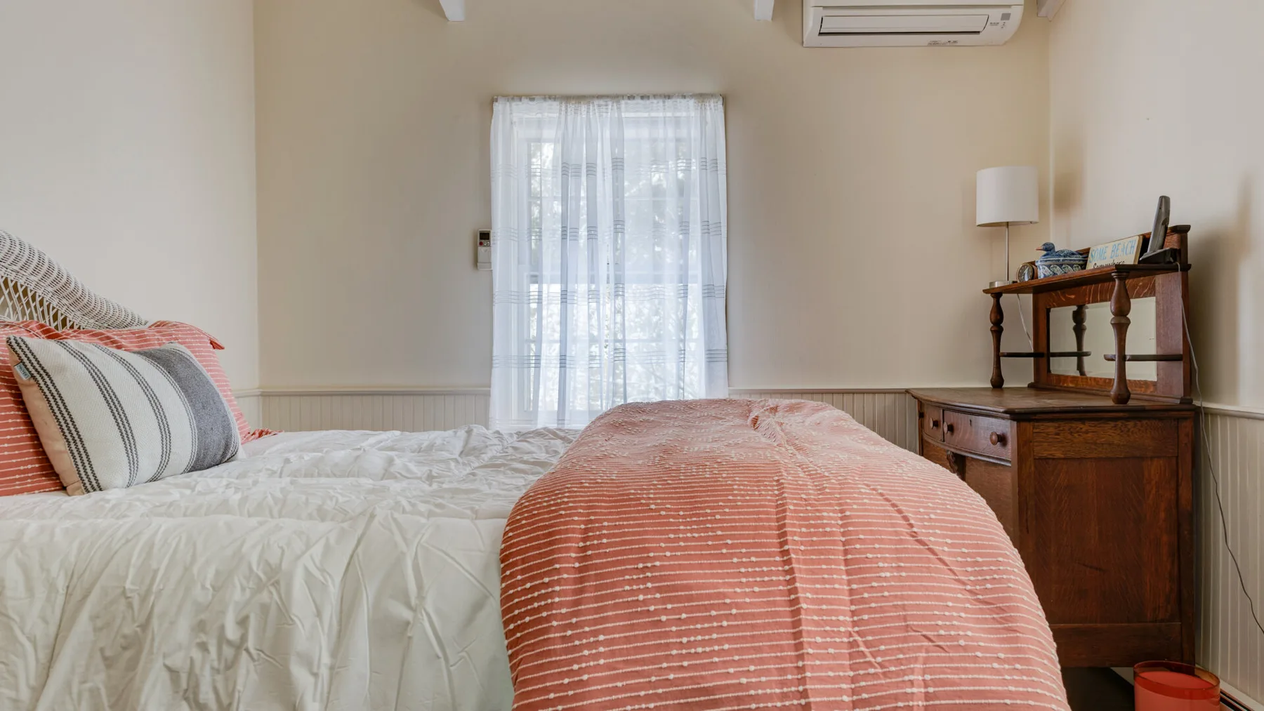 A bedroom with a bed covered in white and coral bedding beside a window.