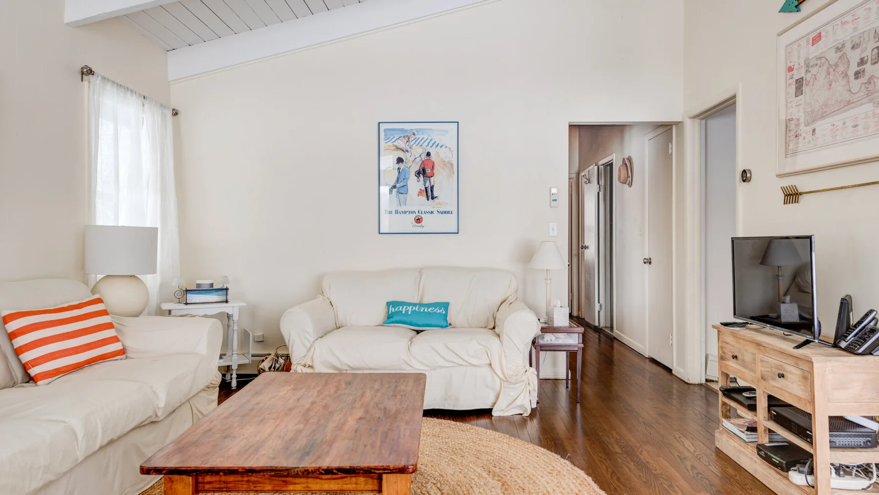 A bright living room with pale seating, a wooden coffee table, and a television.