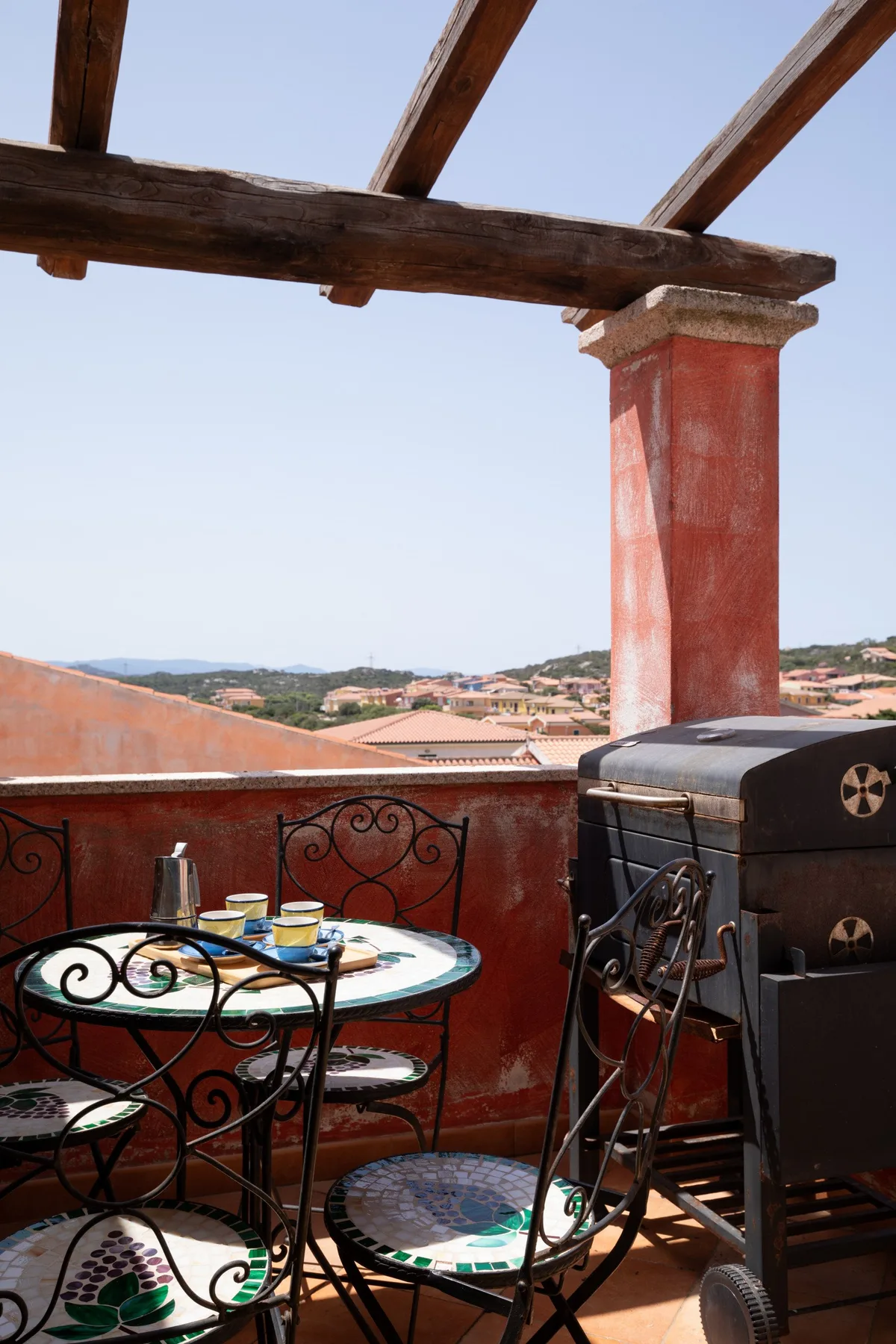 A rooftop terrace with metal seating and a barbecue beneath wooden beams.