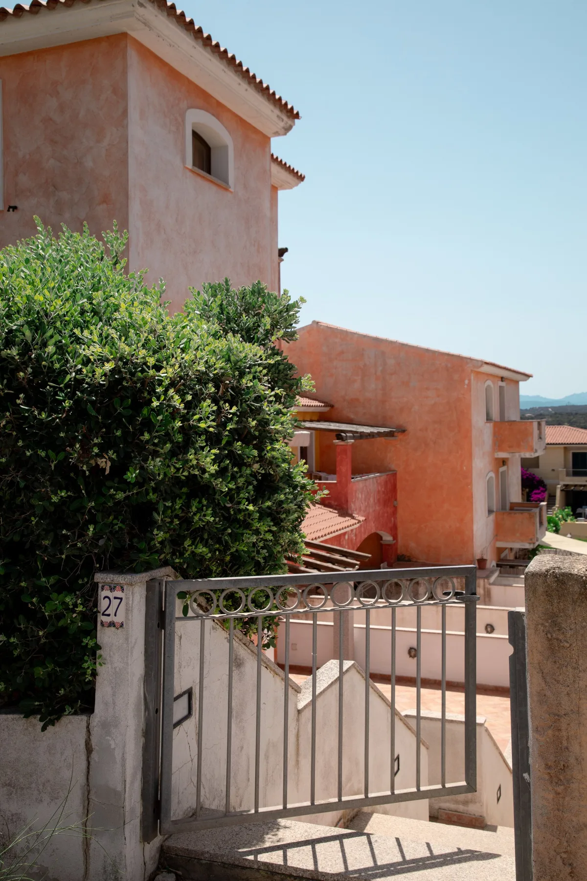 An exterior view of a terracotta-colored building behind a metal gate and leafy shrubs.