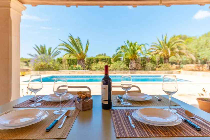 A dining table is set beside a swimming pool, with palm trees beyond.