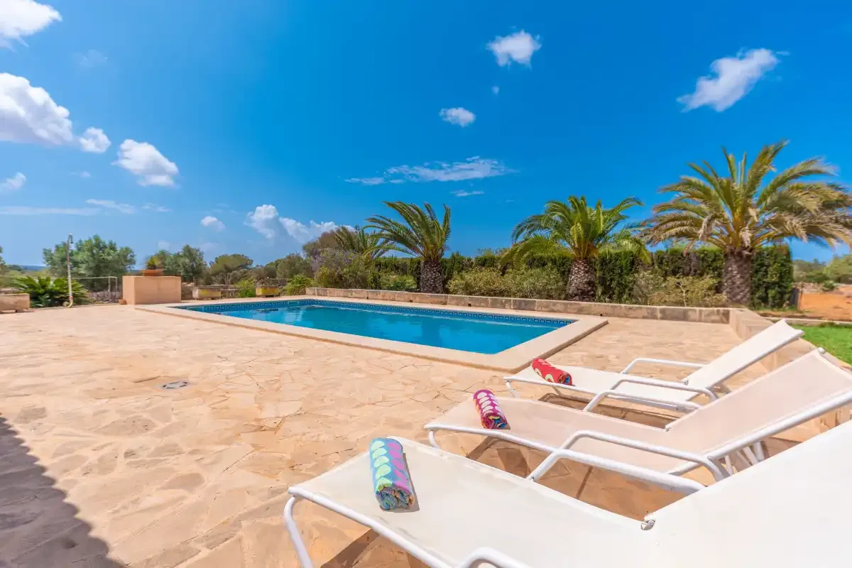An outdoor swimming pool beside a sunlit stone patio and lounge chairs.