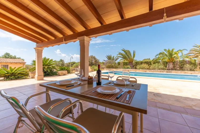 A covered outdoor dining area with a table and chairs overlooks a pool and palm trees.