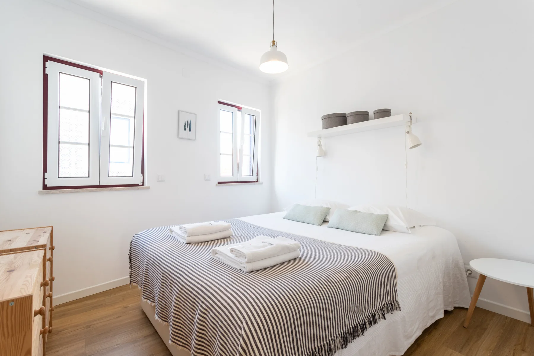 Un dormitorio luminoso con una cama tendida bajo dos ventanas.