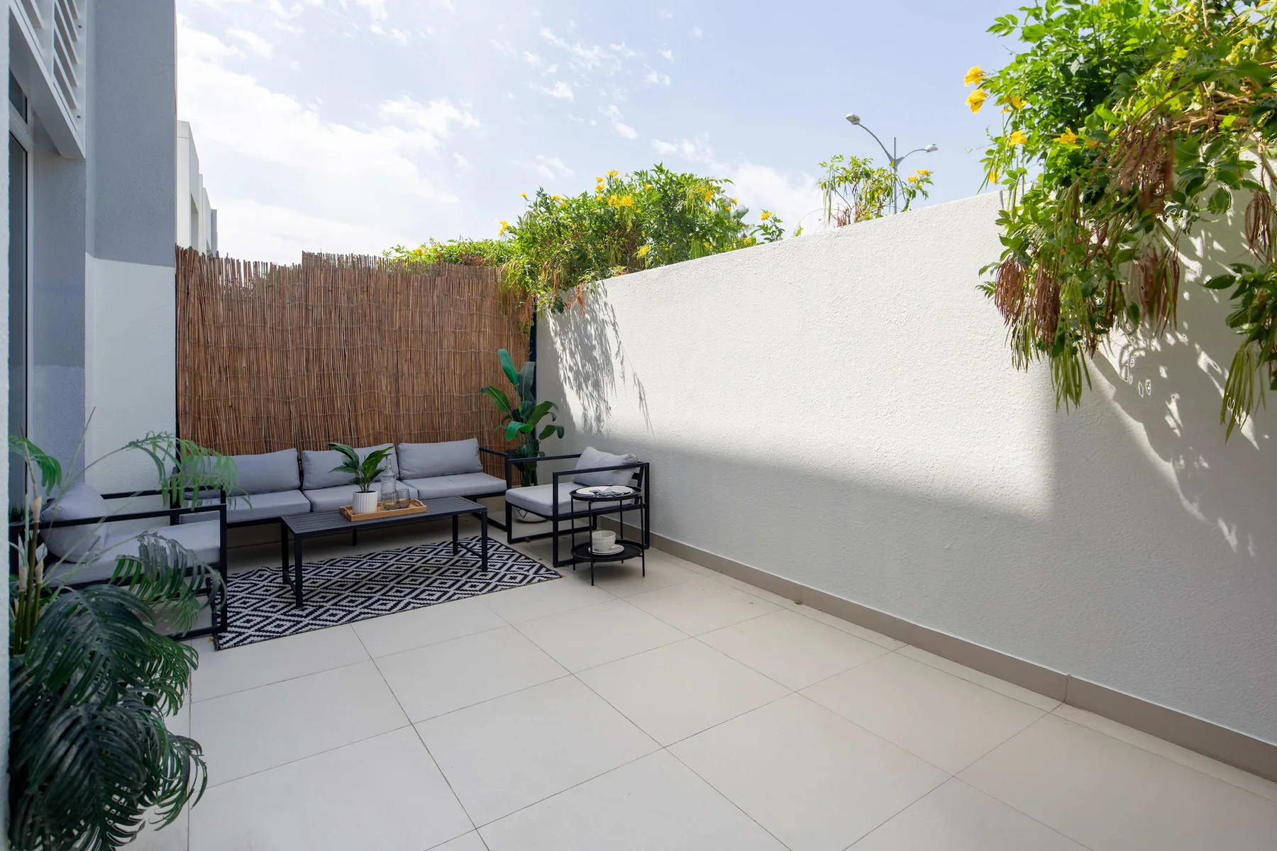 Un patio al aire libre con asientos acolchados, una mesa pequeña y plantas frondosas.