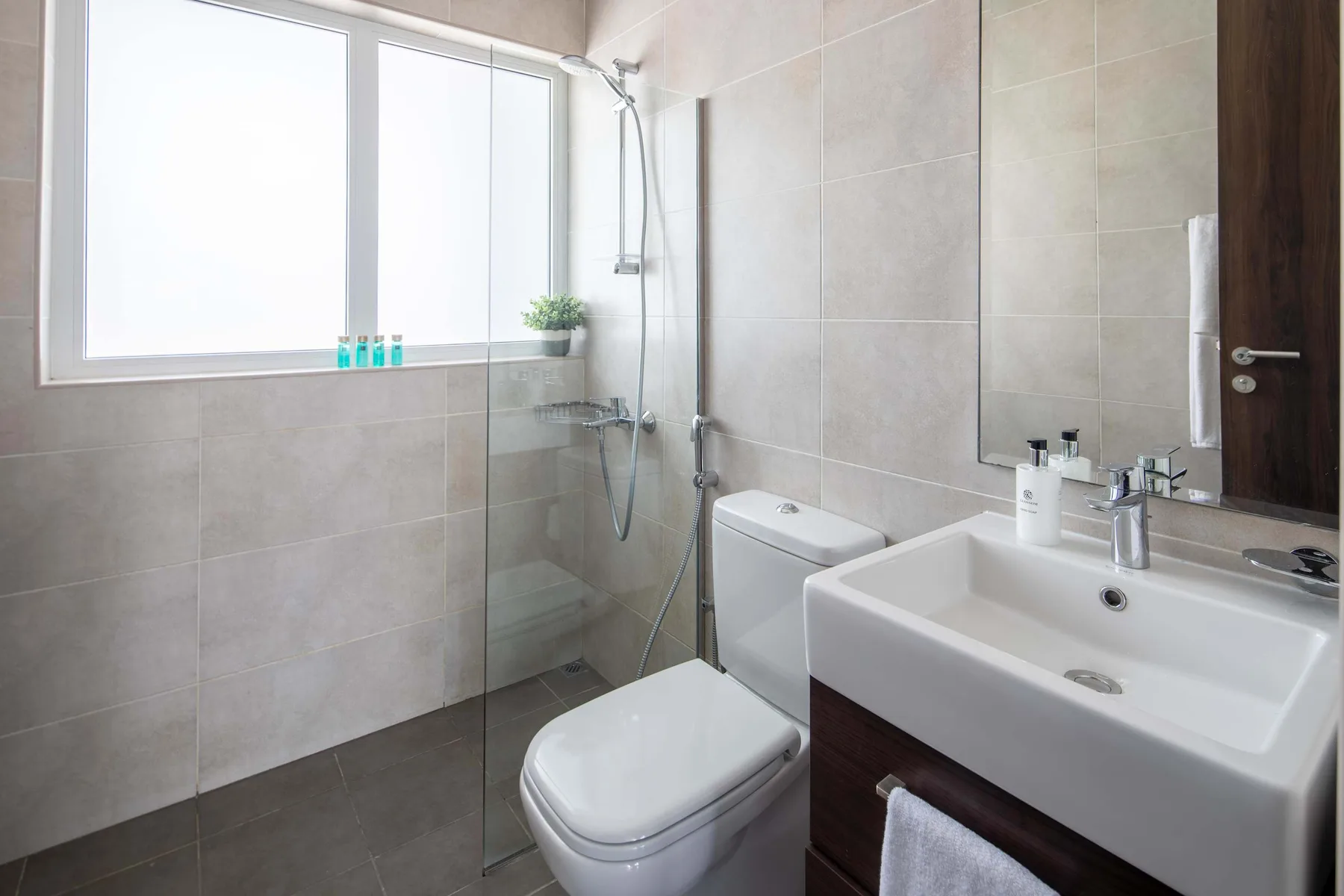 Un baño luminoso con azulejos, ducha, inodoro y lavabo bajo una ventana grande.