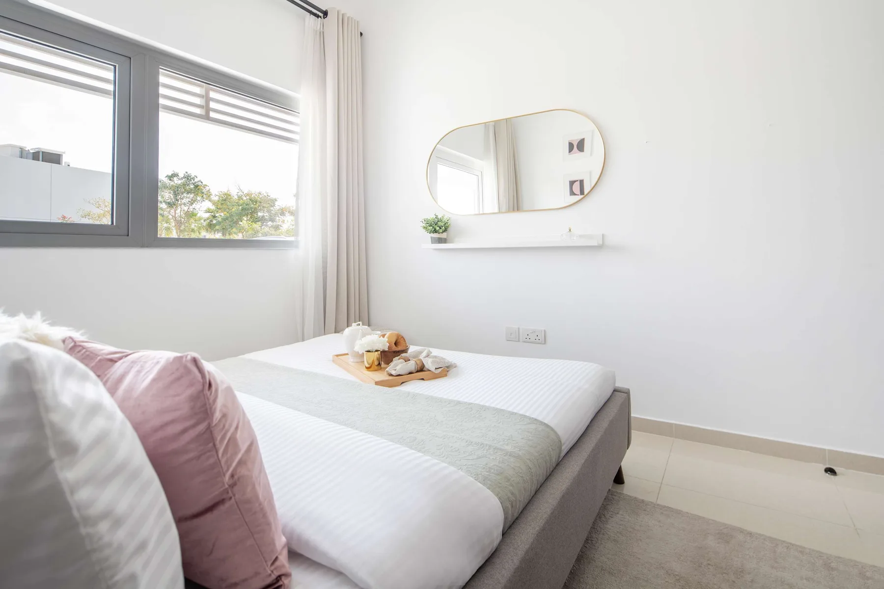 Un dormitorio luminoso con una cama bien tendida, ventanas grandes y un espejo en la pared.