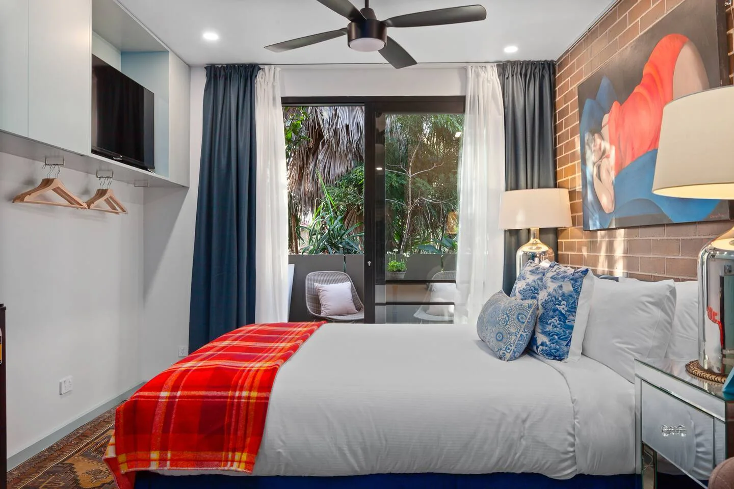 Un dormitorio con una cama bien arreglada, un cuadro colorido y vegetación visible tras las puertas de vidrio.