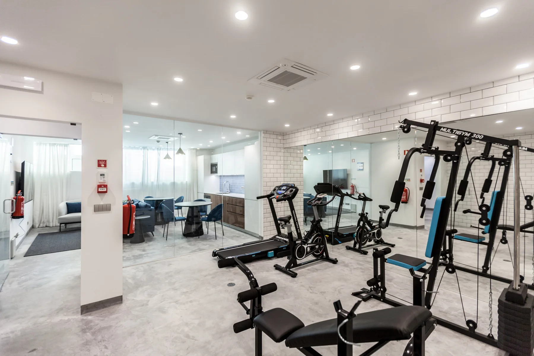 Un gimnasio con máquinas de ejercicio y paredes con espejos junto a un comedor.