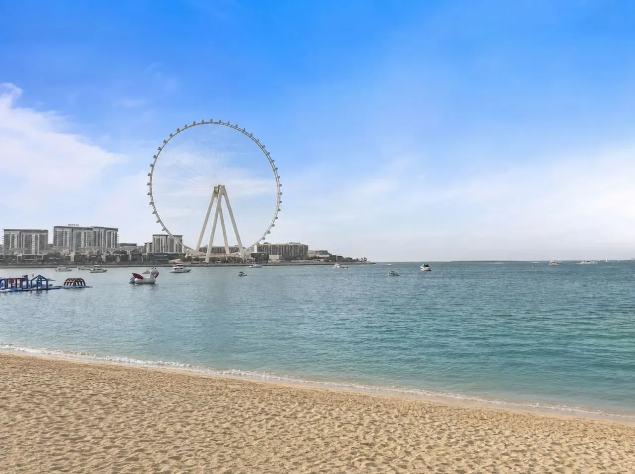A sandy beach borders calm water, with a large observation wheel across the bay.