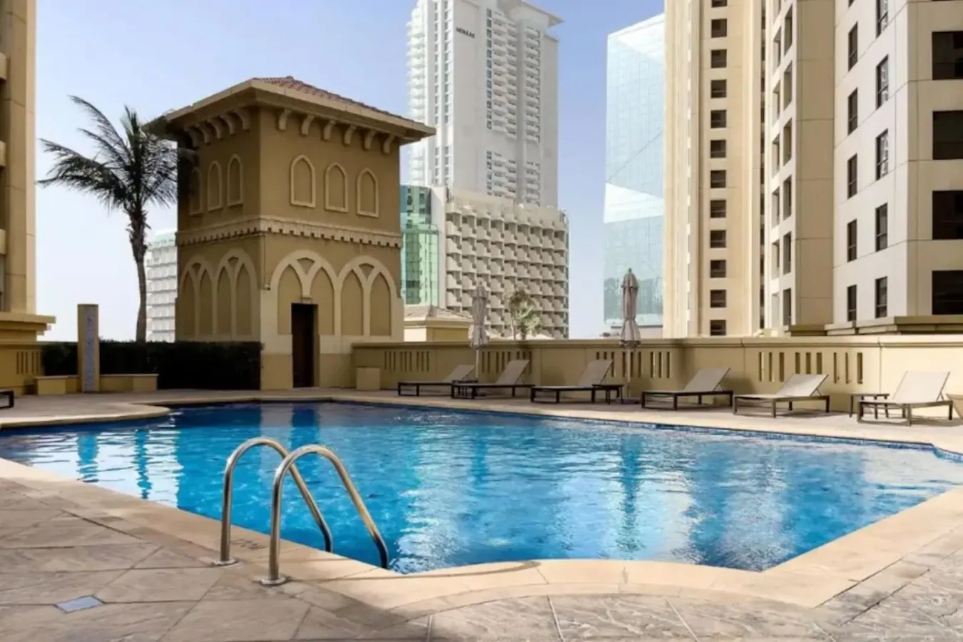 A rooftop swimming pool sits among tall city buildings, with lounge seating along the deck.