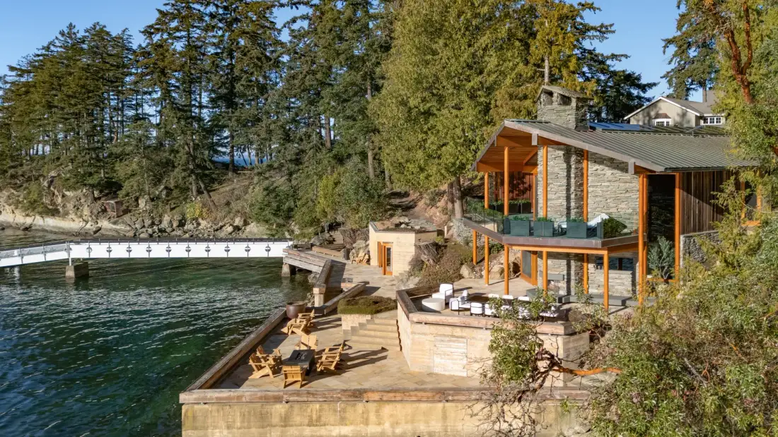A waterfront house with a wooden deck and outdoor seating beside the water.