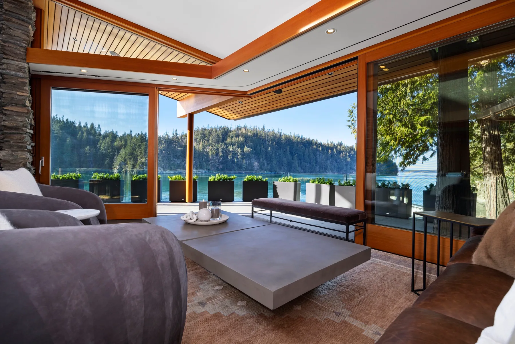 A modern living room opens onto a patio with a view of the water and wooded hills.