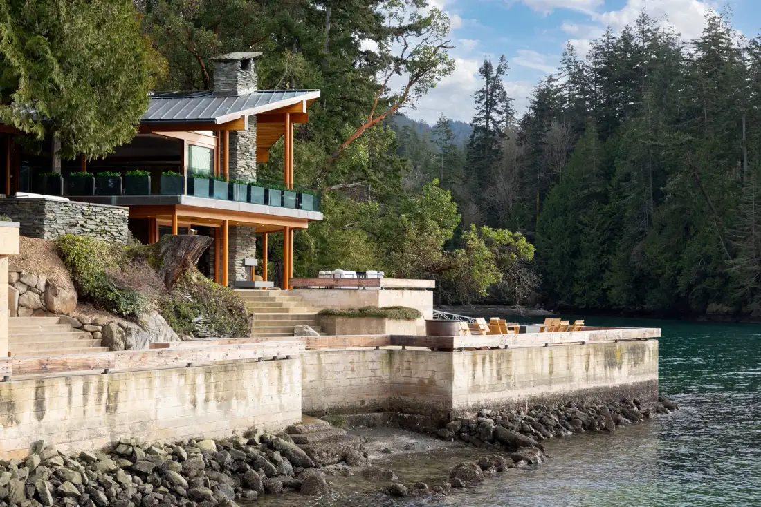 A modern house overlooks a waterfront terrace and wooded shoreline.