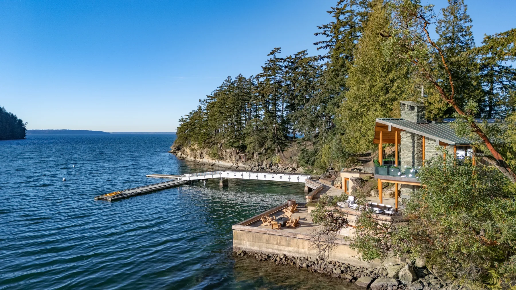 A lakeside cabin sits among trees beside a wooden dock and waterfront terrace.