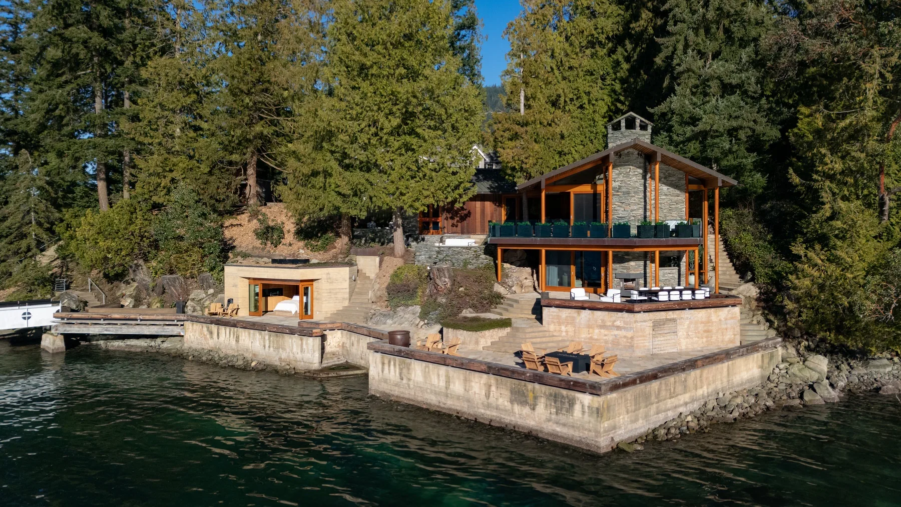 A waterfront house sits beside a stone retaining wall, with trees behind it.