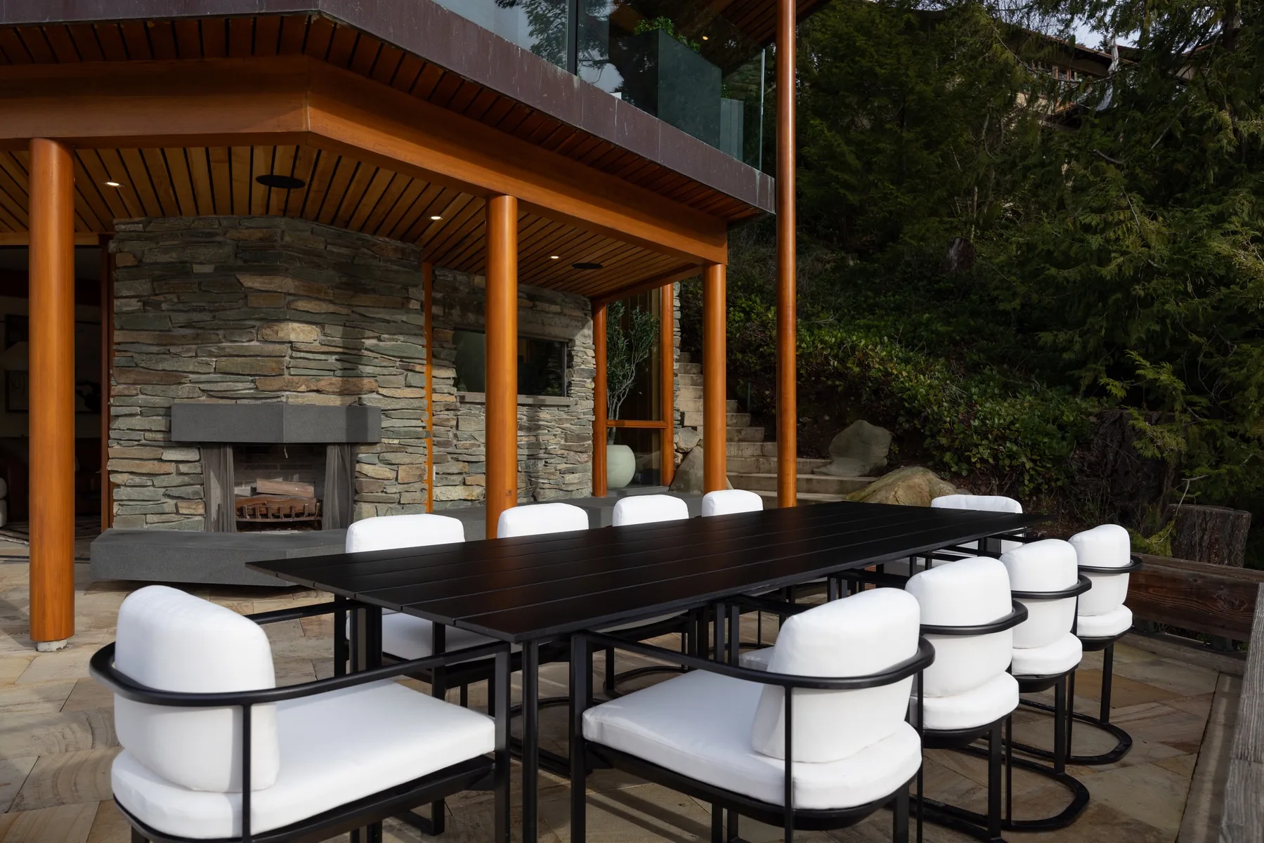 An outdoor dining area with a long table and white seating beside a stone fireplace.
