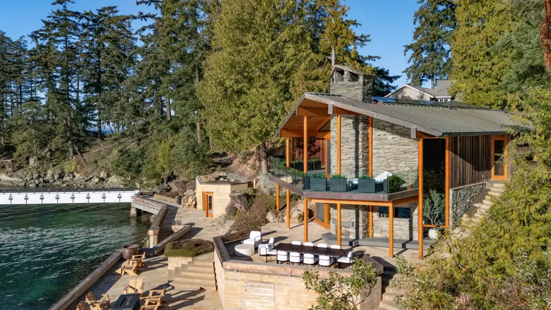 A modern house sits beside a lake, with a terrace and outdoor seating.