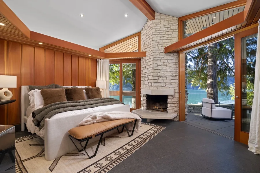A bedroom with a bed, stone fireplace, and large windows overlooking the water.