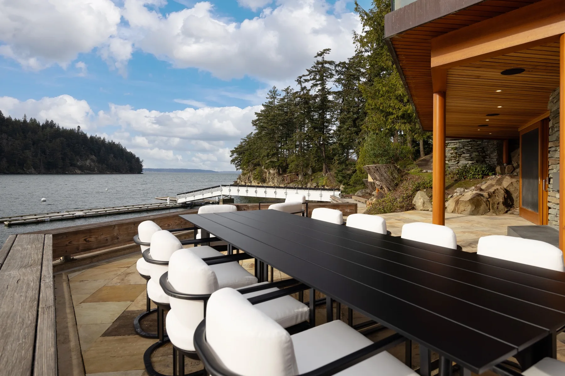 A waterfront dining terrace with a long table and white seating beside the trees.