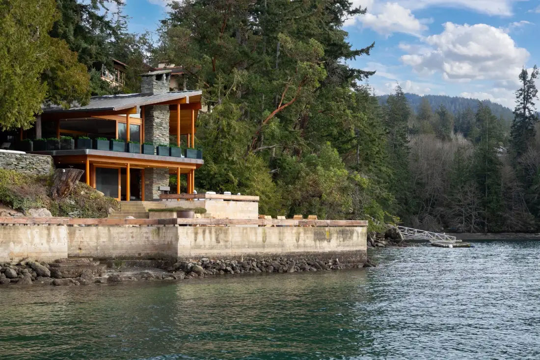 A modern house stands beside a wooded shoreline and calm water.