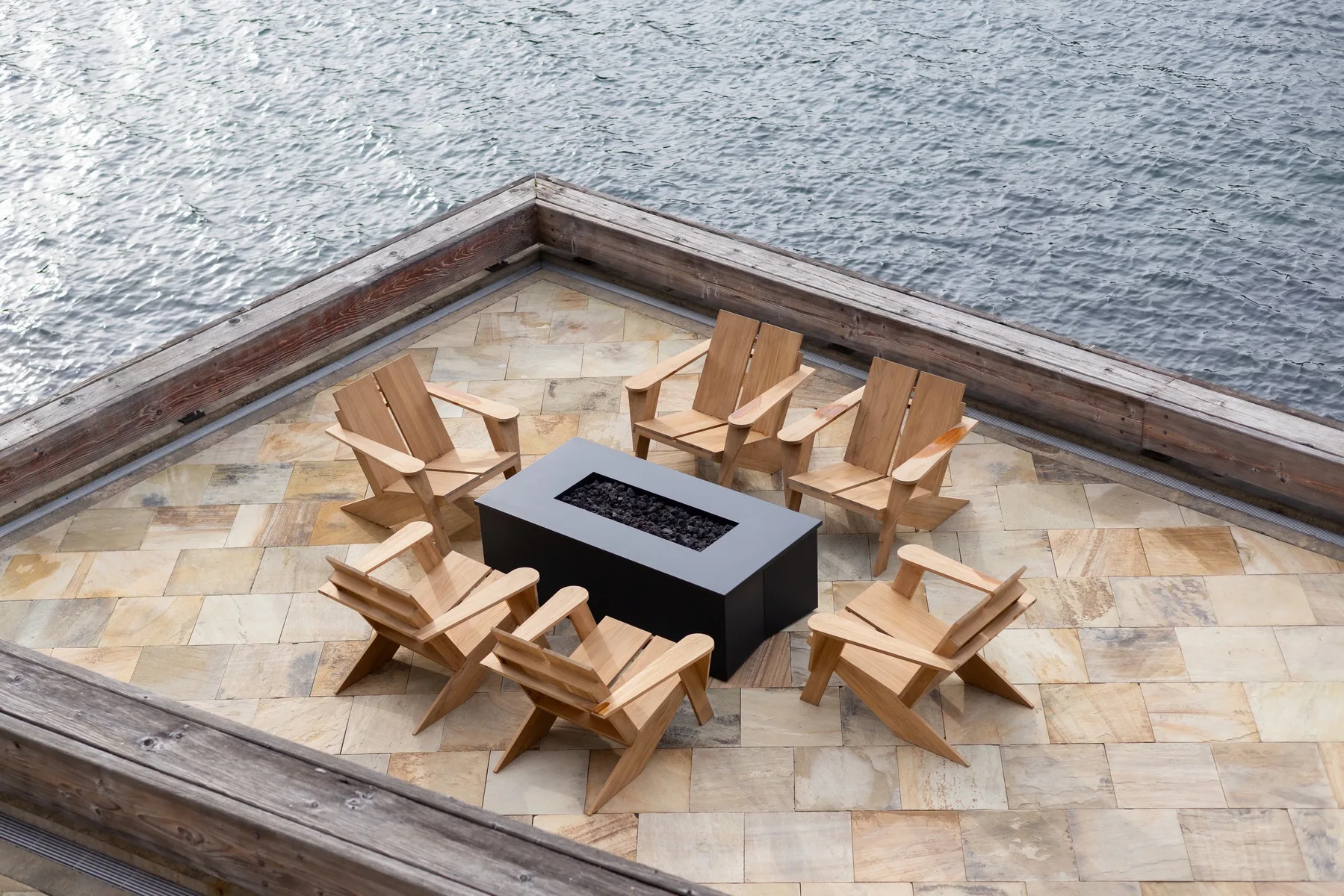 A waterside patio has wooden seating arranged around a rectangular fire feature.